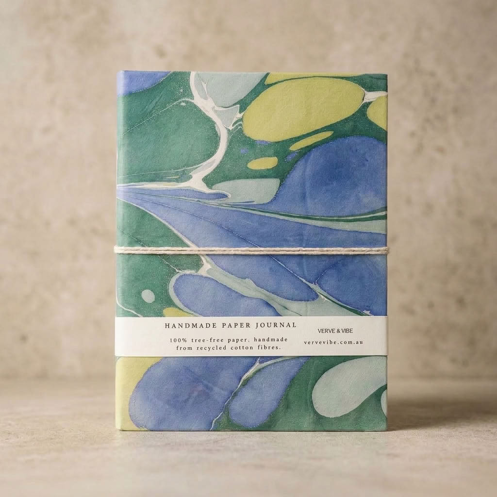 A handmade paper journal wrapped with a twine band, featuring a marbled cover design in shades of blue, green, and yellow, set against a neutral beige background.