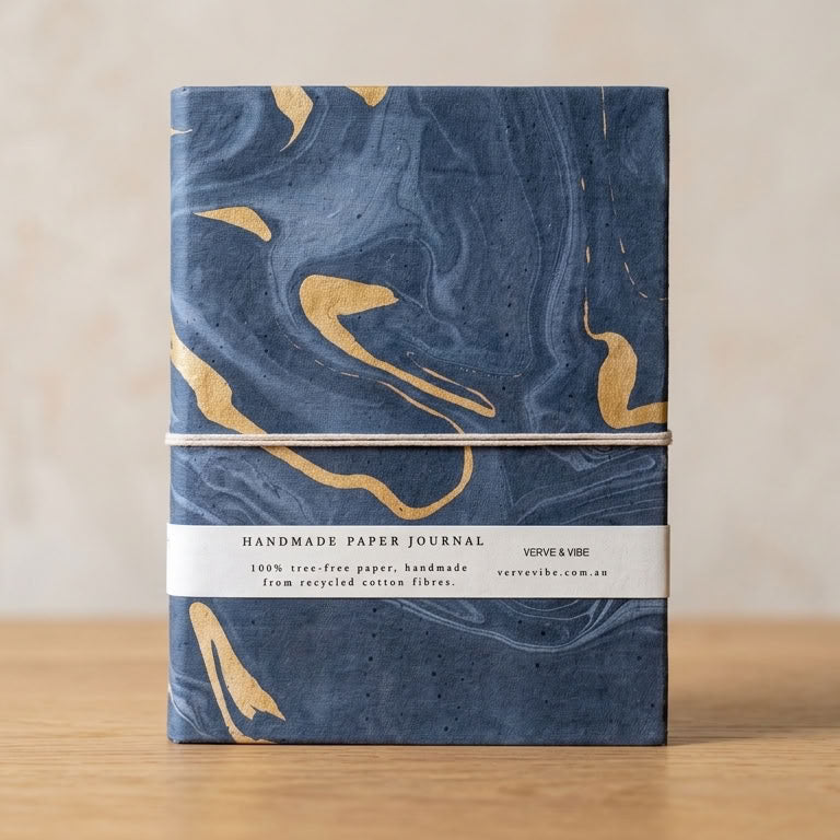 A handmade paper journal with a navy blue and gold marbled cover, wrapped with a white paper band, standing on a light wooden surface against a neutral beige background.