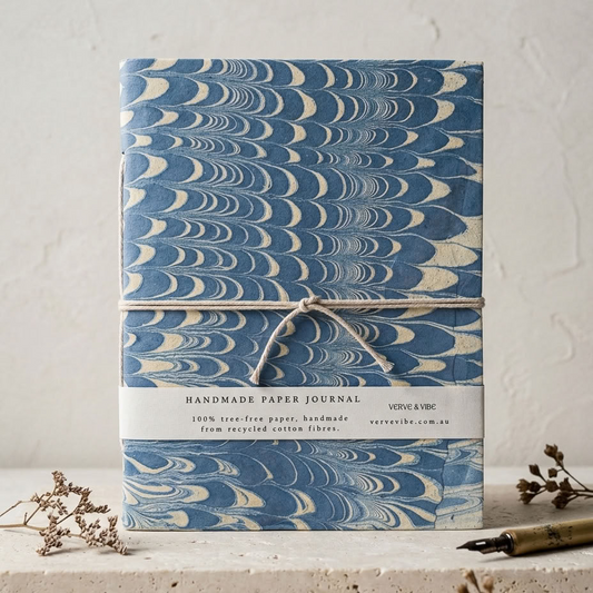A handmade paper journal with a blue and cream marbled wave pattern, wrapped in twine with a white paper label, set on a light stone surface with dried botanicals.