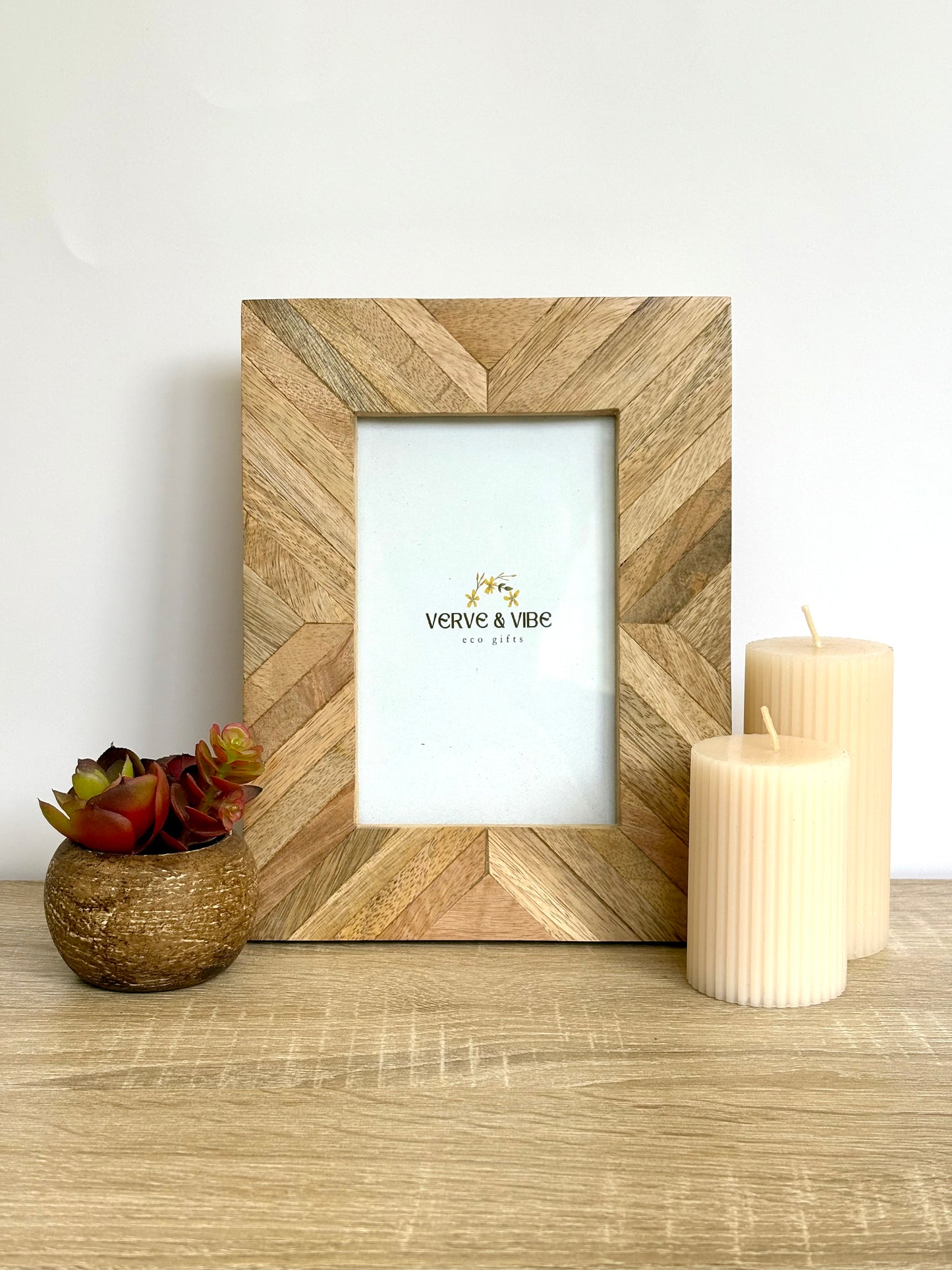 Wooden picture frame with a chevron pattern, placed on a wooden surface with candles and a small plant.