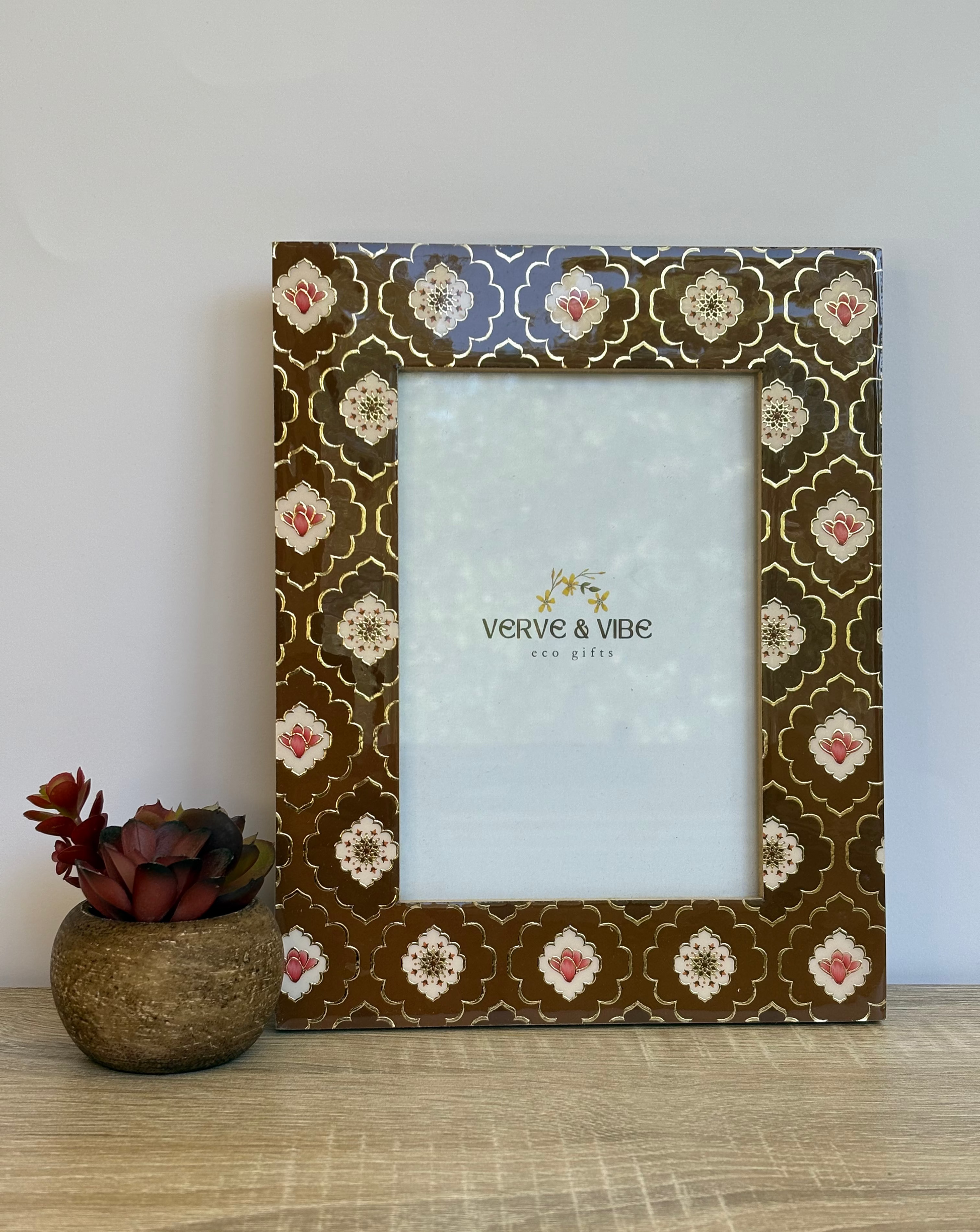 Decorative photo frame with floral pattern next to a small plant on a wooden surface.
