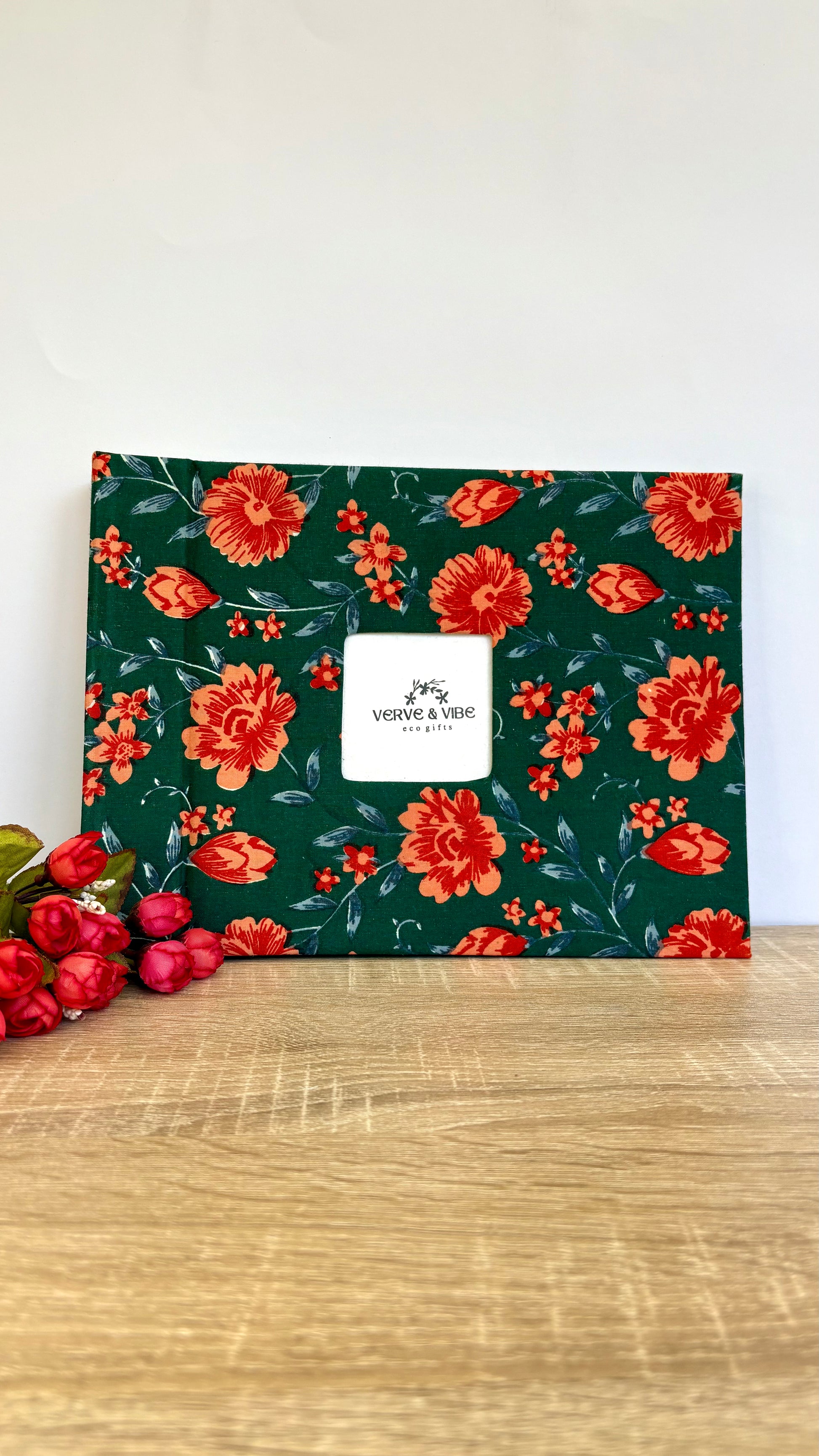 A square fabric gift box or pouch with a dark green background and a red floral pattern, featuring a white central label that reads 'VIRGINIA & NEE'. It is displayed on a light wood surface next to red berries against a plain white wall.